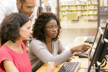 Two adult learners sit in front of a desktop with a teacher, pointing at the screen, the three are deep in thoughtful and open discussion