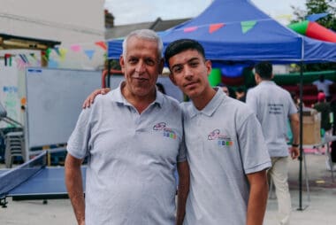 To men sporting our Volunteer shirts pose together One man is older and appears to be a paternal figure for the other man. The other man appears to be a young adult, he has his arm around the shoulders of the older man. The two volunteers have a big smile, and stand in front of bright and colourful bunting hanging above our 'Manor Park Oasis' during launch event