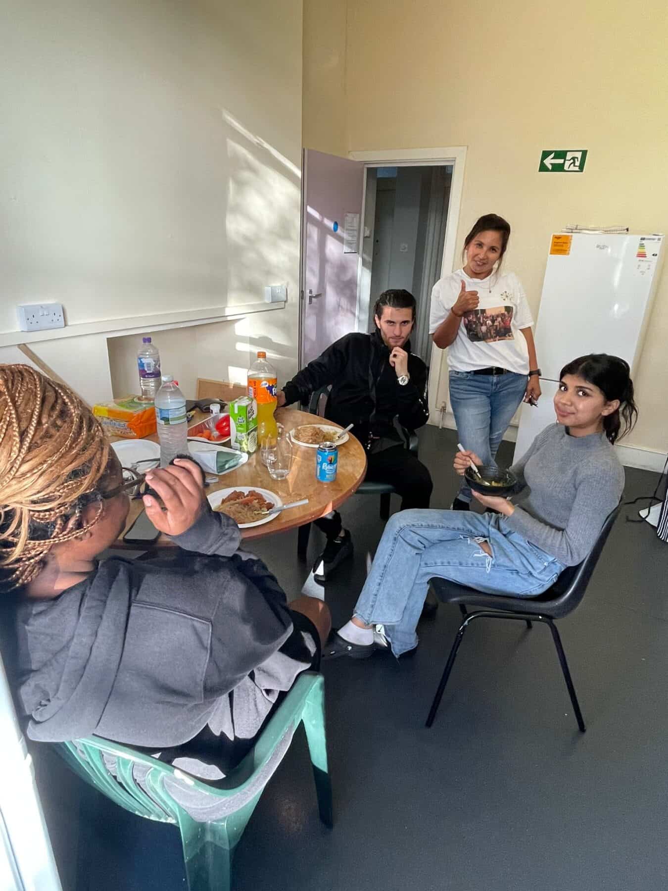 Three residents sit together in supported housing kitchen with Support Worker. The team are sat around a table enjoying a home cooked meal they just made together