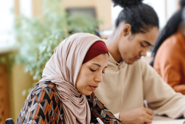 Adult learners sat at a desk, in a warmly lit classroom. A green plant is blurred in the background by a window. Out of focus is a learner in a burnt orange blouse, with a half up curls. next to her is a man in a neutral sweatshirt with a high curly bun (The hair style appears to be a 'Puff Bun'). Next to the man, at the front and in focus is a lady with a burgundy underscarf, and crepe pink hijab. She is staring down at her notebook and attentively making notes